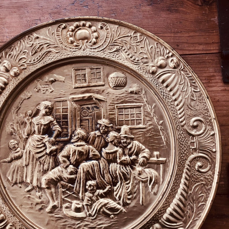 May include: A brass-colored decorative plate with a detailed relief scene of people gathered outside a building. The plate has an ornate floral border and a central scene depicting figures in period clothing. The plate is displayed on a wooden surface.