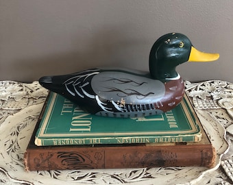 May include: A hand-painted wooden duck decoy with black, green, and brown paint. The duck is sitting on top of two books. The top book is green and has the title "The Londoner" on the spine. The bottom book is brown and has the title "Dorothy Nicholson" on the spine.