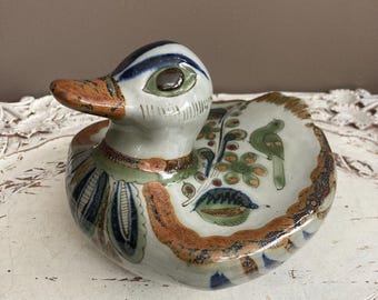 Könnte beinhalten: Handbemalte Keramik-Entenfigur. Die Ente hat einen weißen Körper, einen braunen und orangefarbenen Schnabel sowie blaue und grüne Details. Der Rücken der Ente ist mit einem Muster mit Vögeln und floralen Motiven verziert. Dekoratives Objekt.