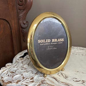 May include: Oval, solid brass picture frame. The frame is hand polished and lacquer coated. The frame is 5" x 7" and has a dark brown backing. The frame has three small feet to stand upright.