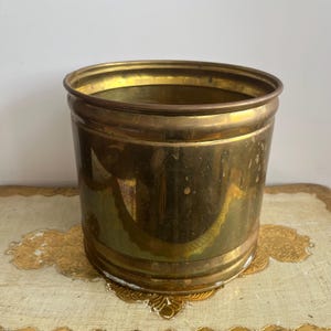 May include: A cylindrical brass planter with a polished metallic finish and a smooth, rounded rim. The exterior surface shows natural variations in colour and texture, reflecting light across its curved body. The vessel is open at the top.