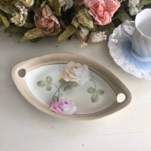 May include: A white oval serving dish with gold trim and a floral design featuring pink and white roses. The dish has two handles on the sides.