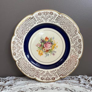 May include: A vintage decorative plate with a scalloped edge and gold trim. The plate features a central floral bouquet with pink roses and yellow tulips, surrounded by a navy blue band and a white background with a gold filigree pattern.