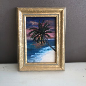 May include: A framed painting of a palm tree on a beach at sunset. The sky is a vibrant pink and purple, and the water is a deep blue. The sand is white and the palm tree is silhouetted against the sky.