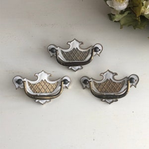 Peut inclure: Trois poignées de tiroir blanches et dorées avec un motif décoratif. Les poignées ont une finition en laiton et un bouton noir de chaque côté.