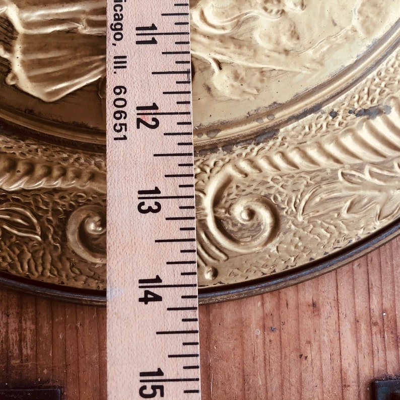 May include: A vintage gold-colored metal decorative plate with embossed floral and scrollwork designs. The plate measures approximately 12 inches in diameter. A wooden ruler is placed vertically along the side of the plate, with the text "Chicago, Ill. 60651" visible.