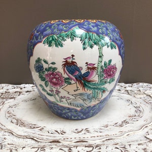 May include: A white ceramic ginger jar with a blue and gold floral design. The jar features two birds with long tails and pink flowers.