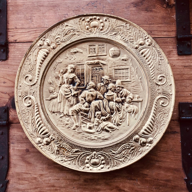 May include: A round, gold-colored metal plate with a detailed relief scene of people gathered outside a building. The plate has an ornate, floral border. The scene depicts several figures, including a woman and children, and men seated at a table.