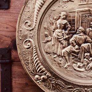May include: A detailed, antique brass plate with a raised relief design. The plate features a central scene of figures in period clothing, surrounded by an ornate, floral border. The plate is set against a wooden background.