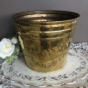 May include: A brass planter with a hammered texture. The planter is cylindrical with a flared rim. The brass has a golden colour. The planter is likely for decorative use, and could hold plants or other items. The diameter is approximately 25 cm.