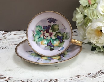 May include: A delicate teacup and saucer set with a floral design. The cup and saucer are white with a gold rim and feature a hand-painted design of purple, yellow, and green flowers.