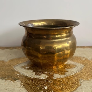 May include: A small, round brass planter with a polished metallic finish and a wide, flared rim. The body of the vessel features subtle vertical indentations around its circumference, creating a soft, faceted appearance with a warm, golden tone.