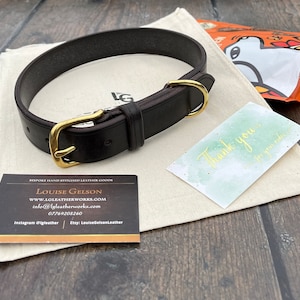 May include: A dark brown leather dog collar with a brass buckle and D-ring. The collar is lying on a white fabric surface with a business card and a thank you note.