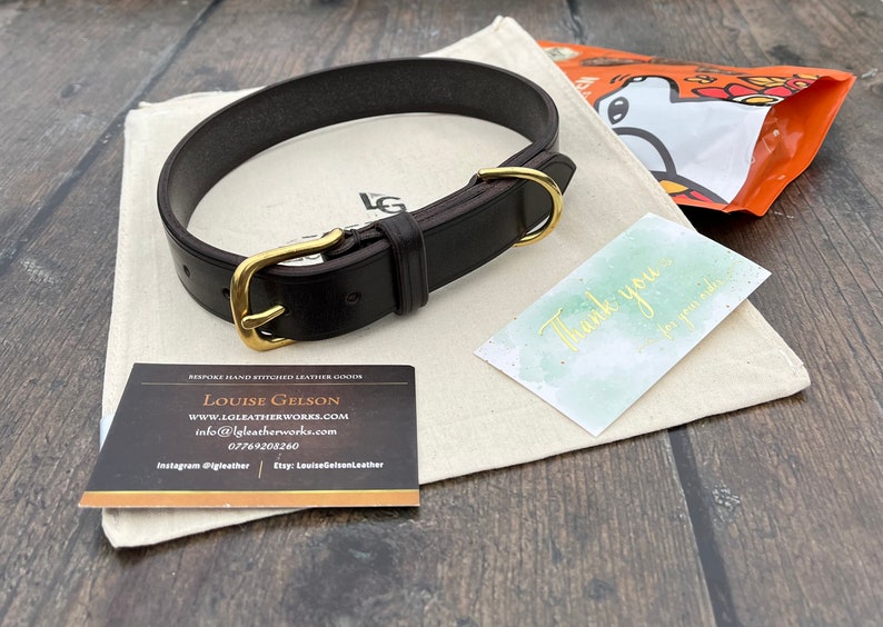 May include: A dark brown leather dog collar with a brass buckle and D-ring. The collar is lying on a white fabric surface with a business card and a thank you note.