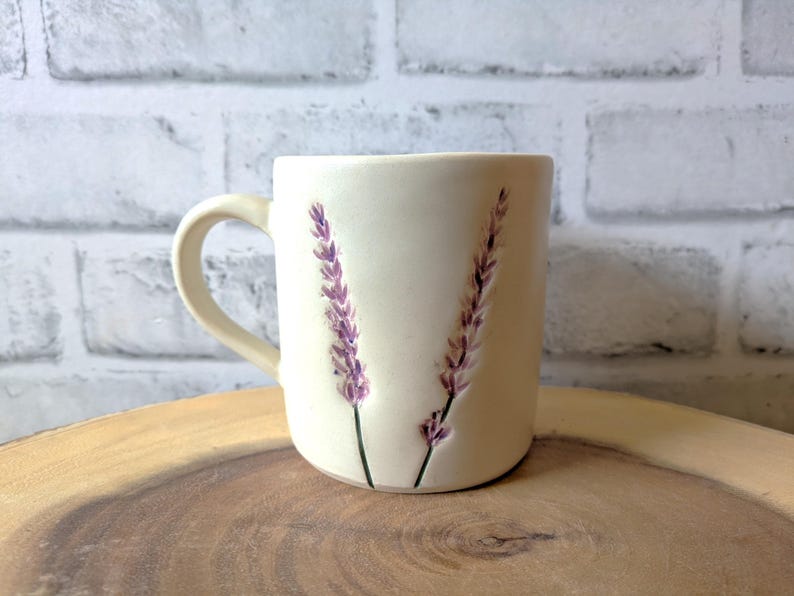 May include: A white ceramic mug with a lavender design. The mug has a handle and is sitting on a wooden surface.