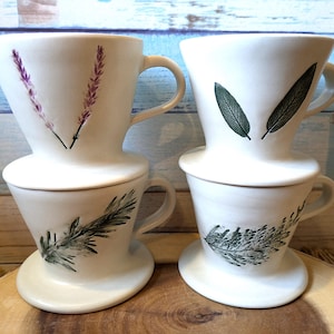 May include: Four white ceramic coffee pour-over drippers with different botanical designs. Two drippers have a lavender design, one has a fern design, and one has a sage design.
