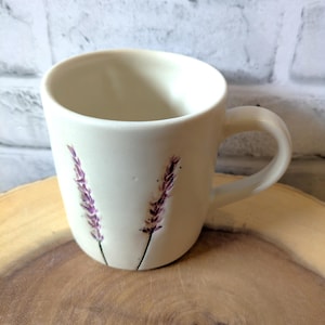 May include: White ceramic mug with a lavender flower design. The mug has a handle and is sitting on a wooden surface.