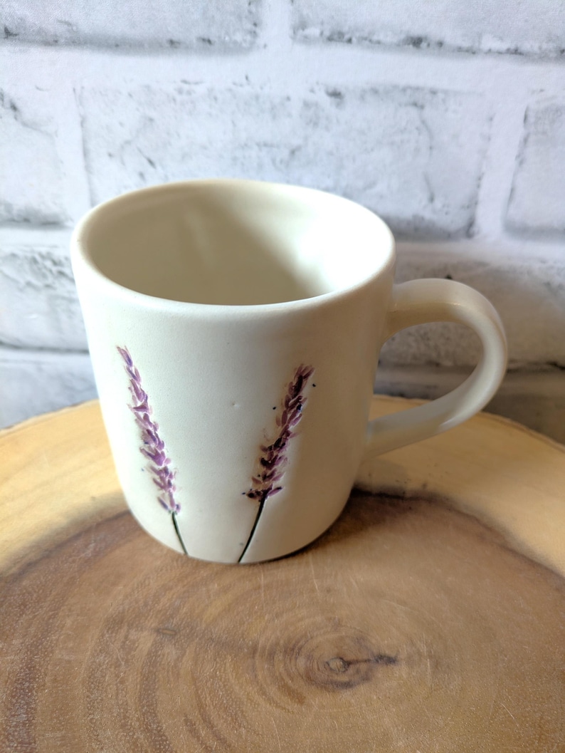 May include: White ceramic mug with a lavender flower design. The mug has a handle and is sitting on a wooden surface.