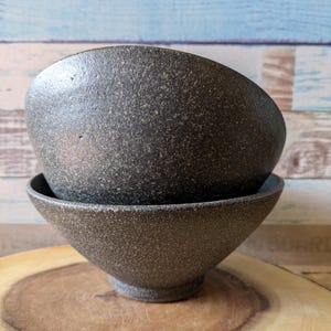 May include: Two dark gray ceramic bowls stacked on top of each other. The bowls have a speckled texture and are sitting on a wooden surface.