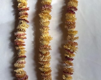 May include: Three strands of dried flowers in shades of yellow, orange, and burgundy. The flowers are arranged in a garland-like pattern, with each individual bloom visible. The background is a plain white surface.