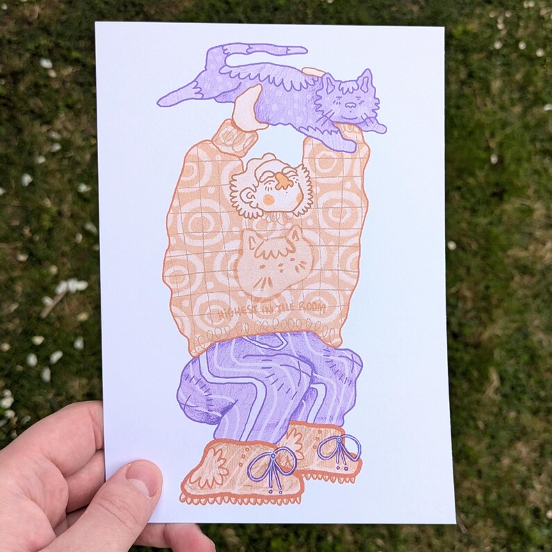 Op de afbeelding: Illustratie van een persoon die een paarse kat boven zijn hoofd houdt. De persoon draagt een oranje trui met een kattenafbeelding en de tekst "HIGHEST IN THE ROOM". Ze dragen ook een paarse broek en oranje laarzen.