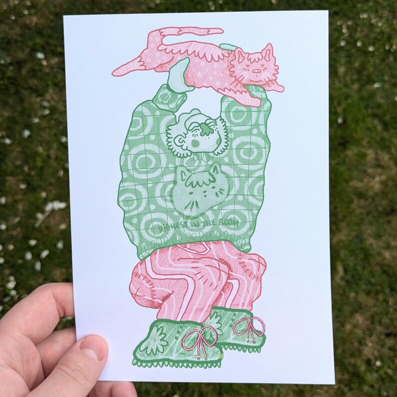 Op de afbeelding: Illustratie van een persoon die een roze kat boven zijn hoofd houdt. De persoon draagt een groene trui met een kattenafbeelding en de tekst "HIGHEST IN THE ROOM". Ze draagt ook een roze broek en groene schoenen. Het kunstwerk staat op wit papier.