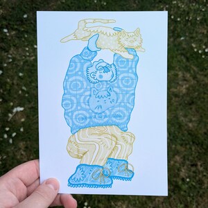 Op de afbeelding: Illustratie van een persoon die een kat boven zijn hoofd houdt. De persoon draagt een blauwe trui met een kattenafbeelding en de tekst "HIGHEST IN THE ROOM". De persoon draagt ook een beige broek en blauwe laarzen. De kat is beige.