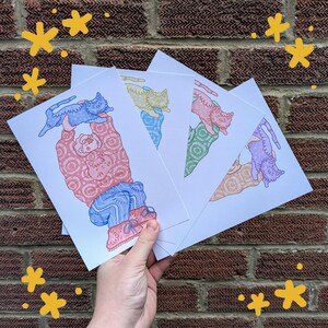 Op de afbeelding: Een hand houdt een stapel kunstprints vast met een grillige illustratie van een persoon die een kat vasthoudt. De persoon draagt een roze trui en een blauwe broek. De katten zijn in blauw, geel, roze en paars. Gele ster krabbels staan op de achtergrond.