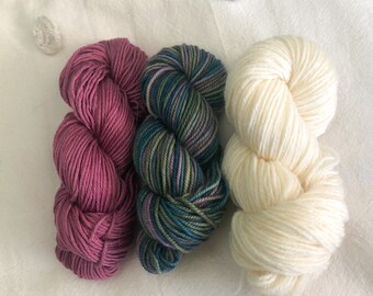 May include: Three skeins of yarn in different colors: a deep pink, a teal and green variegated yarn, and a creamy white.