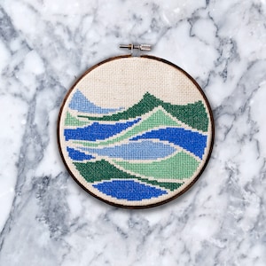 May include: A circular embroidery hoop with a cross-stitch design of abstract blue and green waves on a beige fabric background.