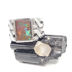 May include: A silver ring with a rectangular opal stone displaying brown, green, and blue colours. The ring sits on a black tourmaline crystal with a clear quartz crystal. The ring has a unique, geometric design.
