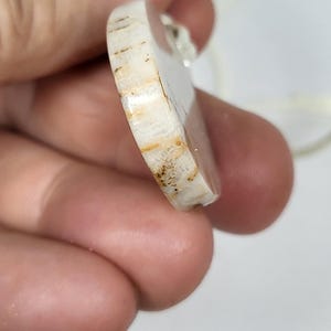Puede incluir: Primer plano de un colgante ovalado blanco y marr&oacute;n con una superficie lisa y pulida. El colgante tiene un patr&oacute;n de veta de madera natural y est&aacute; unido a un cord&oacute;n blanco.