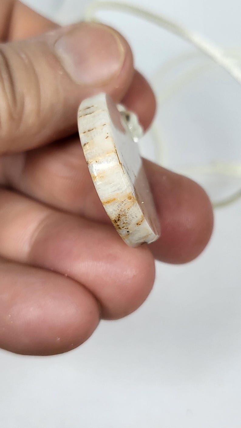 Puede incluir: Primer plano de un colgante ovalado blanco y marr&oacute;n con una superficie lisa y pulida. El colgante tiene un patr&oacute;n de veta de madera natural y est&aacute; unido a un cord&oacute;n blanco.