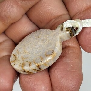 Puede incluir: Un colgante ovalado blanco y marr&oacute;n con un cord&oacute;n blanco anudado. El colgante tiene un aspecto natural y org&aacute;nico con vetas y remolinos marrones.