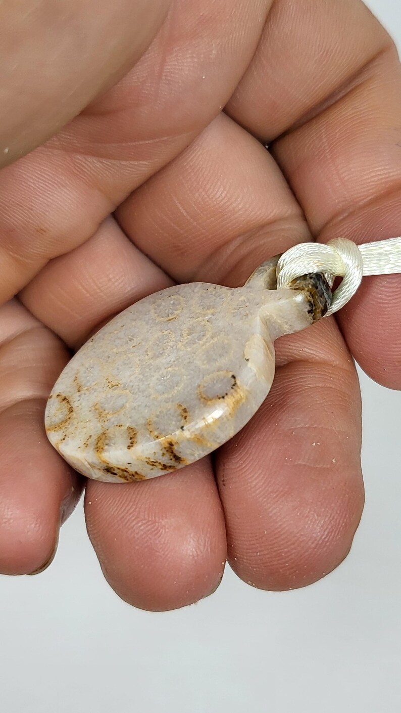 Puede incluir: Un colgante ovalado blanco y marr&oacute;n con un cord&oacute;n blanco anudado. El colgante tiene un aspecto natural y org&aacute;nico con vetas y remolinos marrones.
