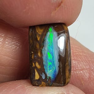 May include: A rectangular boulder opal gemstone featuring a dark brown ironstone matrix with a central, elongated vein of vibrant, iridescent green and blue play-of-color that exhibits a bright, reflective luster across its polished surface.