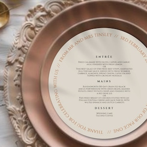 May include: A wedding table setting featuring a blush pink plate, ornate charger, and gold-coloured cutlery. A menu with the text "From Mr and Mrs Tinsley" and "3rd February 2024" is placed on the plate.