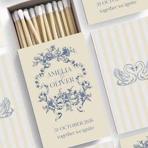 May include: A box of matches with a cream-colored background and a blue floral design. The box is open, revealing the matchsticks with white tips. The box is decorated with the names "Amelia & Oliver" and the date "31 October 2026".