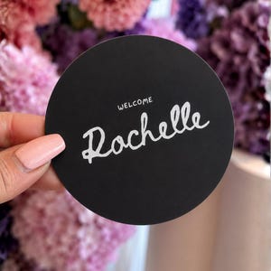 May include: A circular black sign featuring the white text "WELCOME" in a small sans-serif font above the larger, stylised white script text "Rochelle". The sign has a smooth, matte finish and a clean, rounded edge, presenting a simple design.