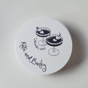 May include: A stack of white, circular coasters featuring a black and white illustration of two cocktail glasses, each with a heart garnish. The text "Katie and Bailey" and "7 November 2025" are also printed on the coasters.