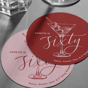 May include: Two round drink coasters, one deep red and one light pink, each featuring a white line drawing of a martini glass with olives. The text reads "ANNETTE IS sixty" and "Sassy, Sweet, Sour & F**king Sensational" in a mix of script and sans-serif fonts.