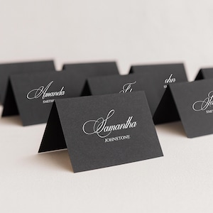 May include: Black place cards with white calligraphy names. The cards are folded and stand upright. The name "Samantha Johnstone" is visible on one card. Other cards have names like "Amanda Smith".