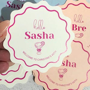May include: A stack of scalloped-edge circular paper coasters in cream, light blue, and peach. Each coaster features the text hello Sasha, a cocktail glass illustration, and the text WELCOME TO CHRISTINE'S 40TH printed in vibrant pink ink.