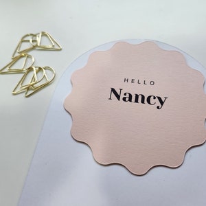 May include: A pink scalloped card with the text "Hello Nancy" in black lettering.