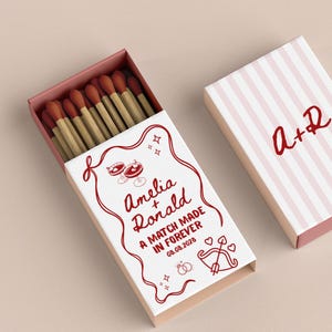 Może przedstawiać: Pudełko zapałek w czerwono-białym wzorze. Pudełko jest otwarte, ukazując kilka zapałek z czerwonymi główkami. Na froncie pudełka widnieją imiona "Amelia + Ronald" oraz napis "A MATCH MADE IN FOREVER 08.08.2028". Widoczne jest drugie pudełko.