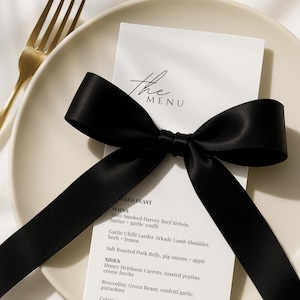 May include: A white menu card, featuring the text "the MENU", is adorned with a black satin ribbon bow. The menu details a selection of dishes, such as slow-smoked beef sirloin and salt-roasted pork belly, on a cream plate with a gold fork.
