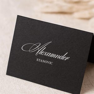 May include: A black place card with the name "Alexander" in elegant white script above the name "Stanovic". The card is angled, revealing a glimpse of the back. The background is a neutral beige.
