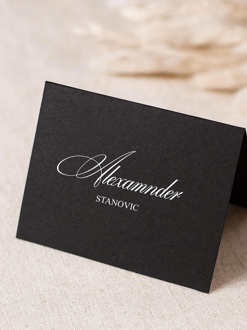 May include: A black place card with the name "Alexander" in elegant white script above the name "Stanovic". The card is angled, revealing a glimpse of the back. The background is a neutral beige.