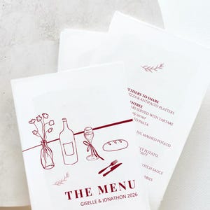 May include: White wedding menu cards with burgundy illustrations of wine, bread, and flowers. The menu includes the text "THE MENU" and the names "Giselle & Jonathon 2026". The menu also lists appetizers and entrees.