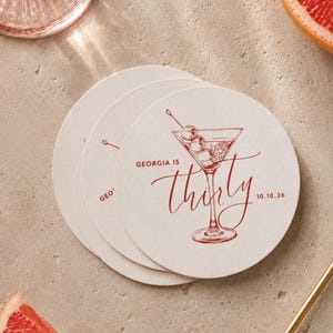 May include: A stack of circular, off-white paper coasters featuring a red ink illustration of a martini glass with three olives. The text reads "GEORGIA IS thirty" and "10.10.26" in a combination of block and script fonts printed on the surface.
